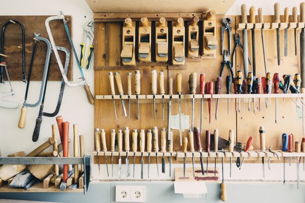 Eine Wand in der Werkstatt ist mit Holz- und Metallwerkzeugen bestückt, darunter Sägen, Hämmer, Meißel, Schraubenzieher und Hobel, die an einer Holzplatte aufgehängt sind.